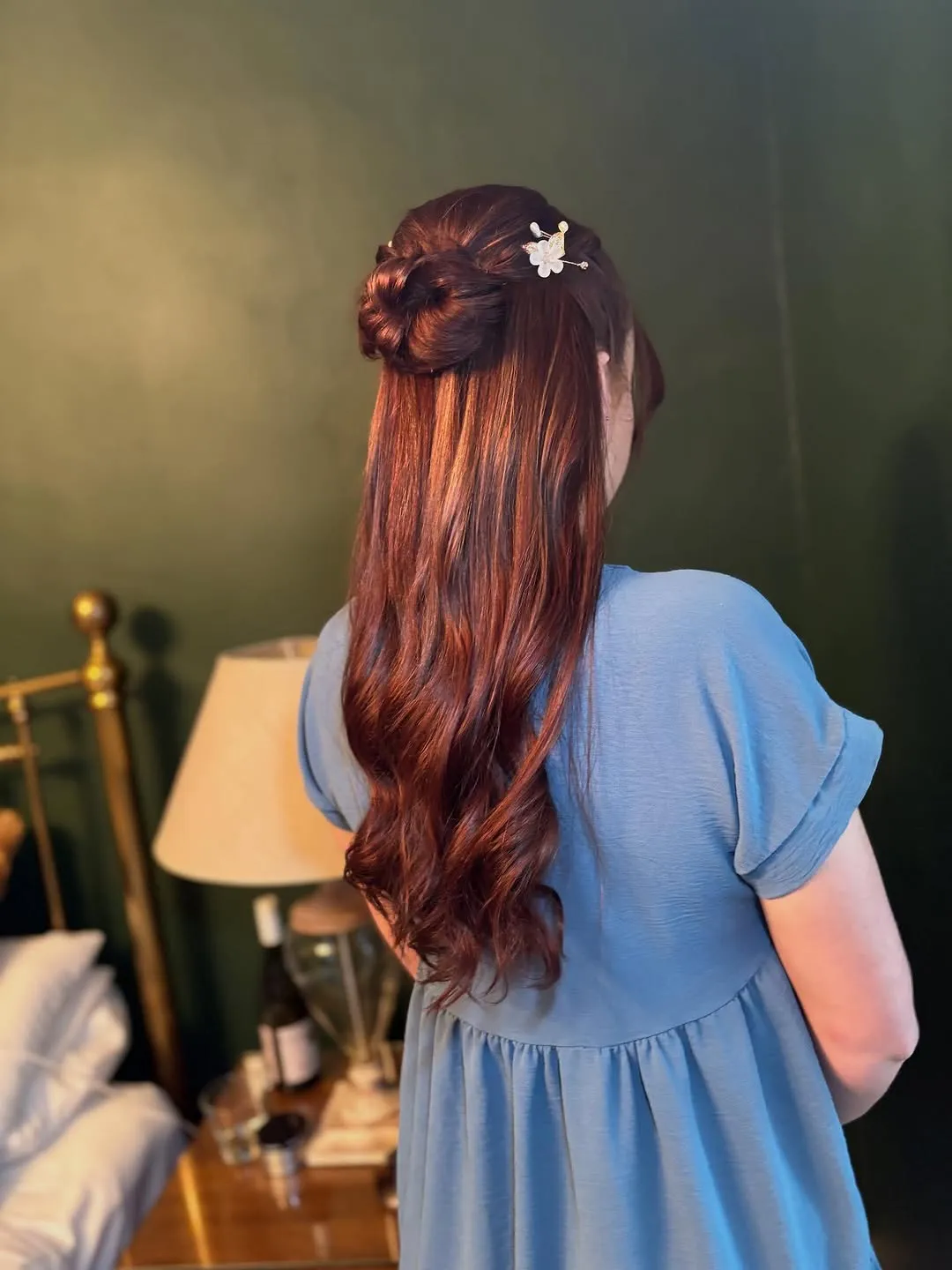 Copper half-up style finished with a small white flower pin, against a deep green wall.