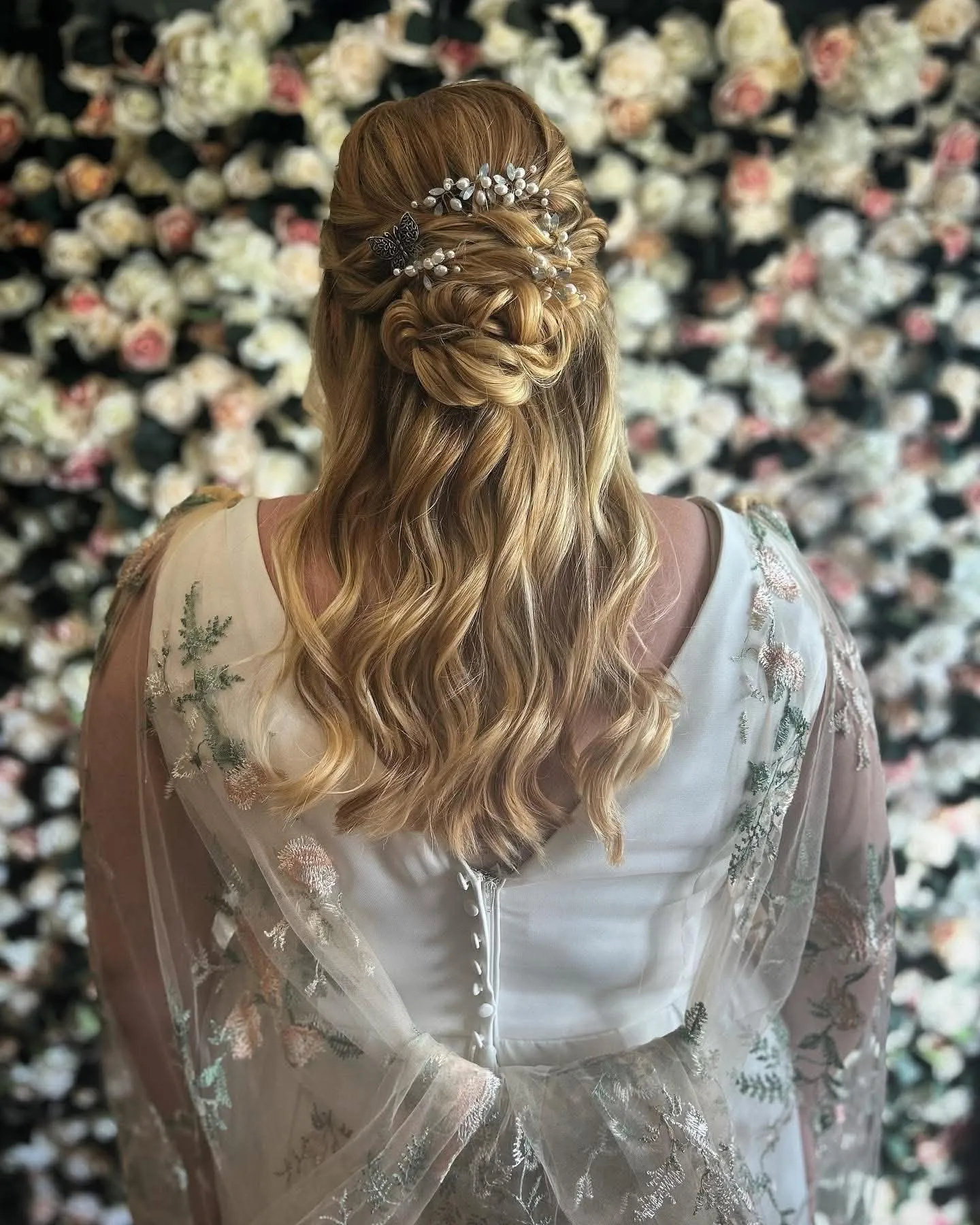 Half-up bridal style with a woven twist, a crystal hair vine and long blonde waves, in front of a wall of flowers