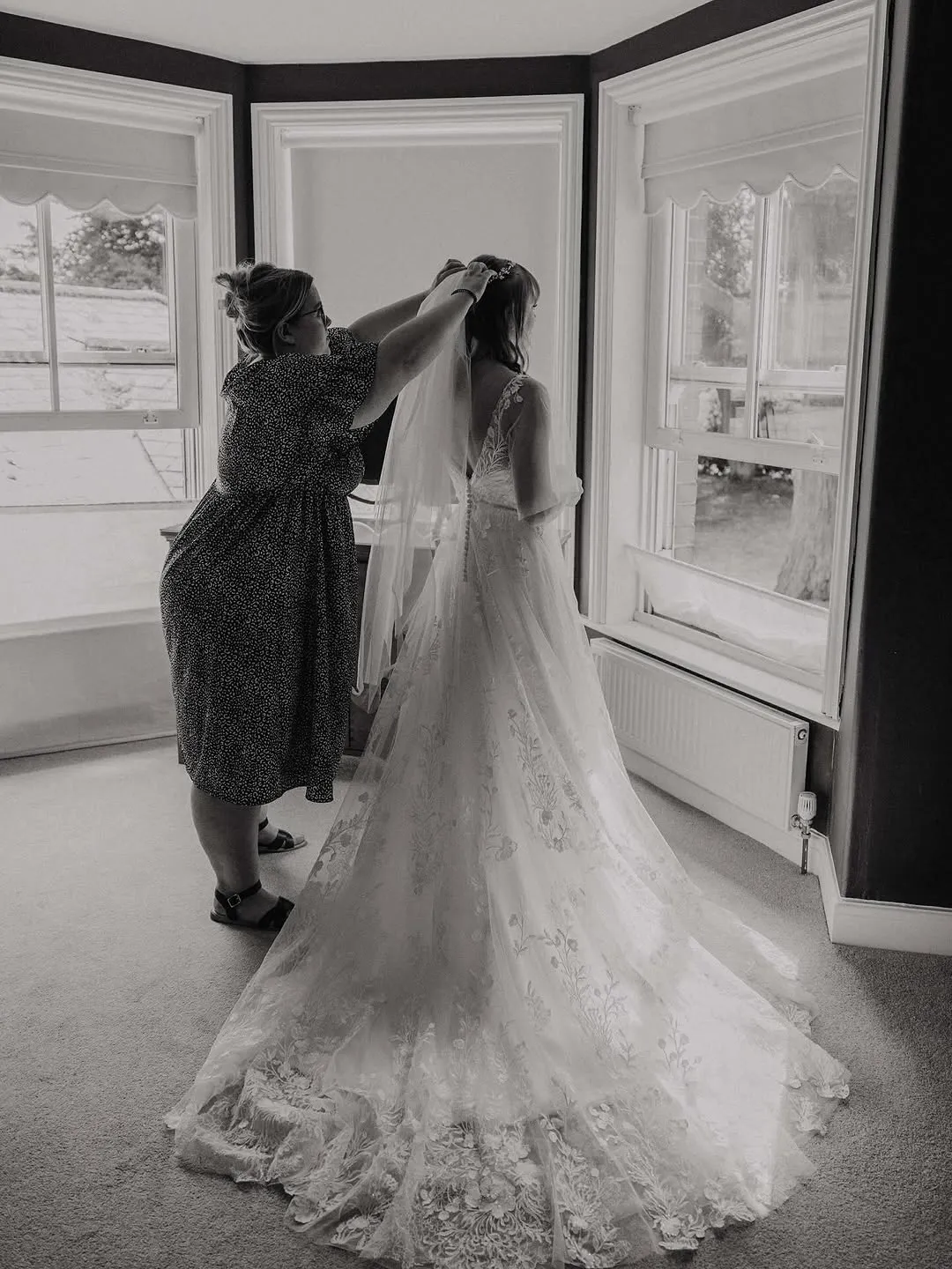 Rosie lifting a veil onto a bride in a lace gown, standing in a bright bay window.